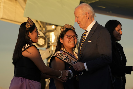 President Joe Biden appearing in the background at a campaign event, symbolizing his subtle presence in the 2024 election race